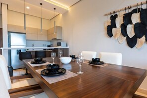 A modern dining area with a wooden table set for four, a kitchen in the background, and a wall with decorative leaf patterns. - Luxury 2 BR PH w/Private Pool, Walk to Beach (Tulum)