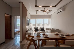 A modern dining area with a wooden table set for four, a ceiling fan, and a large window providing a view of the outdoors. - Luxury 2BR w/Rooftop Pool & Gym I Walk to Beach (Tulum)
