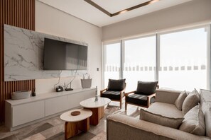 A modern living room with a flat-screen TV mounted on a marble wall, a sofa, two armchairs, a coffee table, and a large window with a view. - Luxury 2BR w/Rooftop Pool & Gym I Walk to Beach (Tulum)