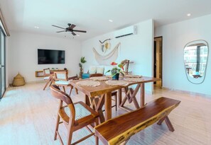 A dining area with a wooden table, chairs, and a bench. There is a ceiling fan, a flat-screen TV mounted on the wall, and a hammock decoration. - Relaxing 2BR Oasis w/ Pool & Balcony | Sleeps 6 (Tulum)