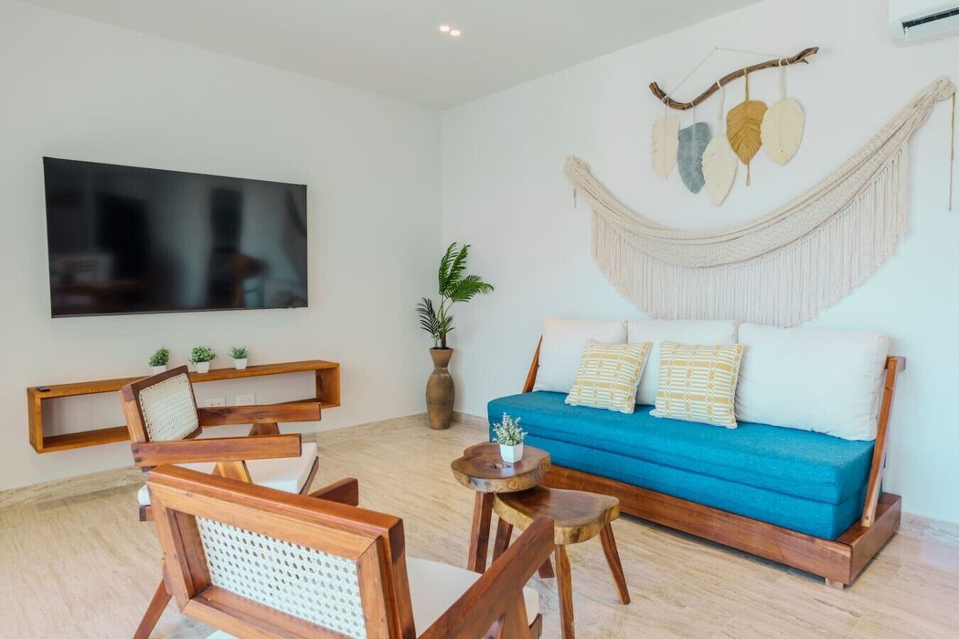 A modern living room with a blue sofa, wooden chairs, a TV, and a macramé wall hanging.