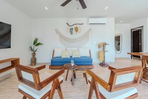 A modern living room with a blue sofa, wooden chairs, a round coffee table, and a hammock. - Relaxing 2BR Oasis w/ Pool & Balcony | Sleeps 6 (Tulum)