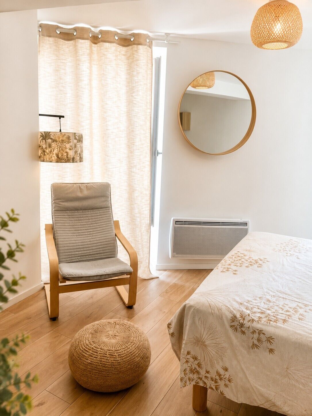 A bedroom with a bed, a chair, a round mirror, a wall-mounted heater, and a window with curtains.