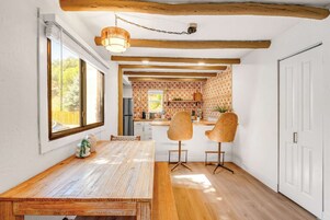 Un comedor con una mesa de madera y dos sillas, una cocina al fondo y una puerta blanca a la derecha.