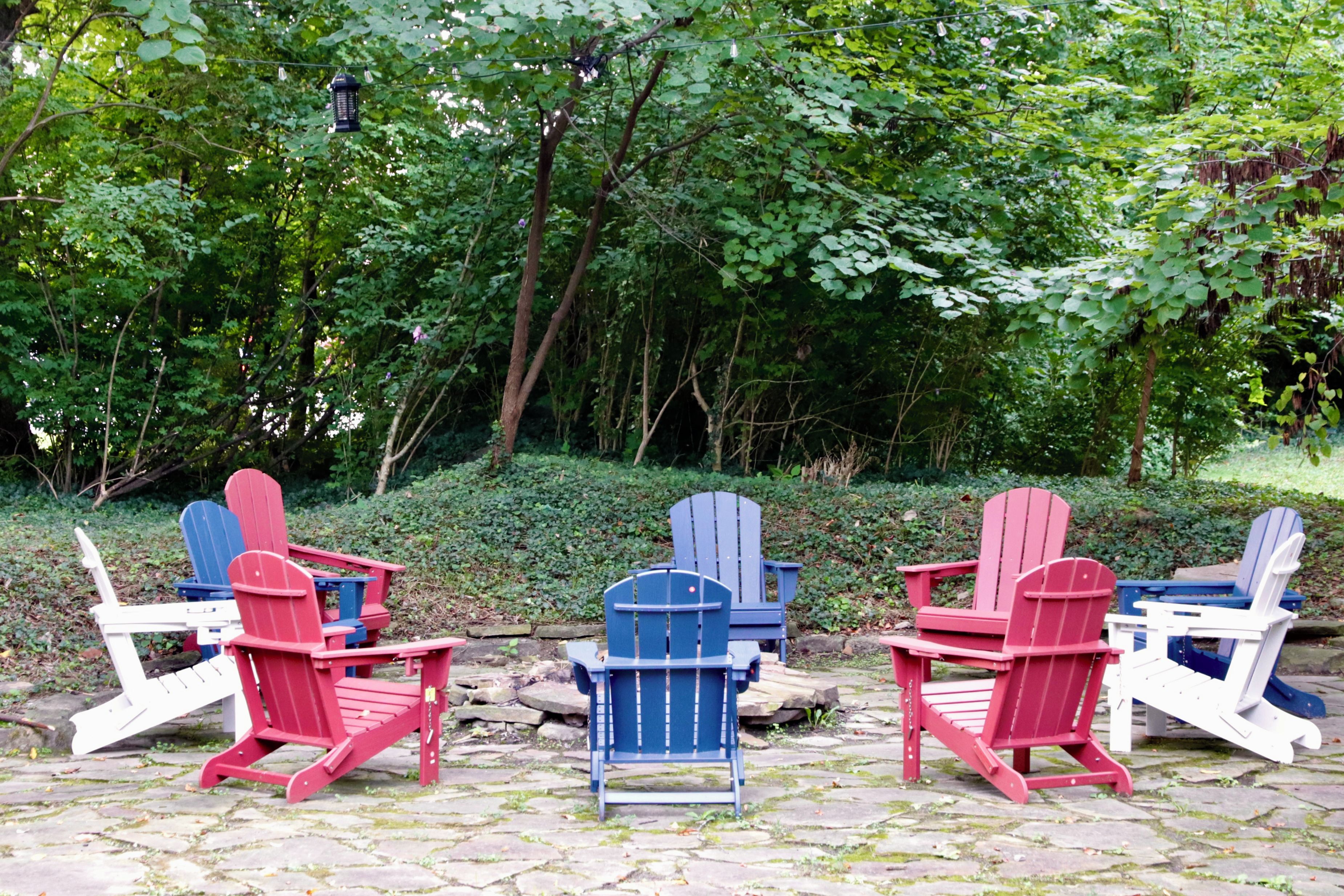 Three Adirondack chairs in red, blue, and white colors, arranged around a stone fire pit in a wooded area.