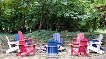 Three Adirondack chairs in red, blue, and white colors, arranged around a stone fire pit in a wooded area.