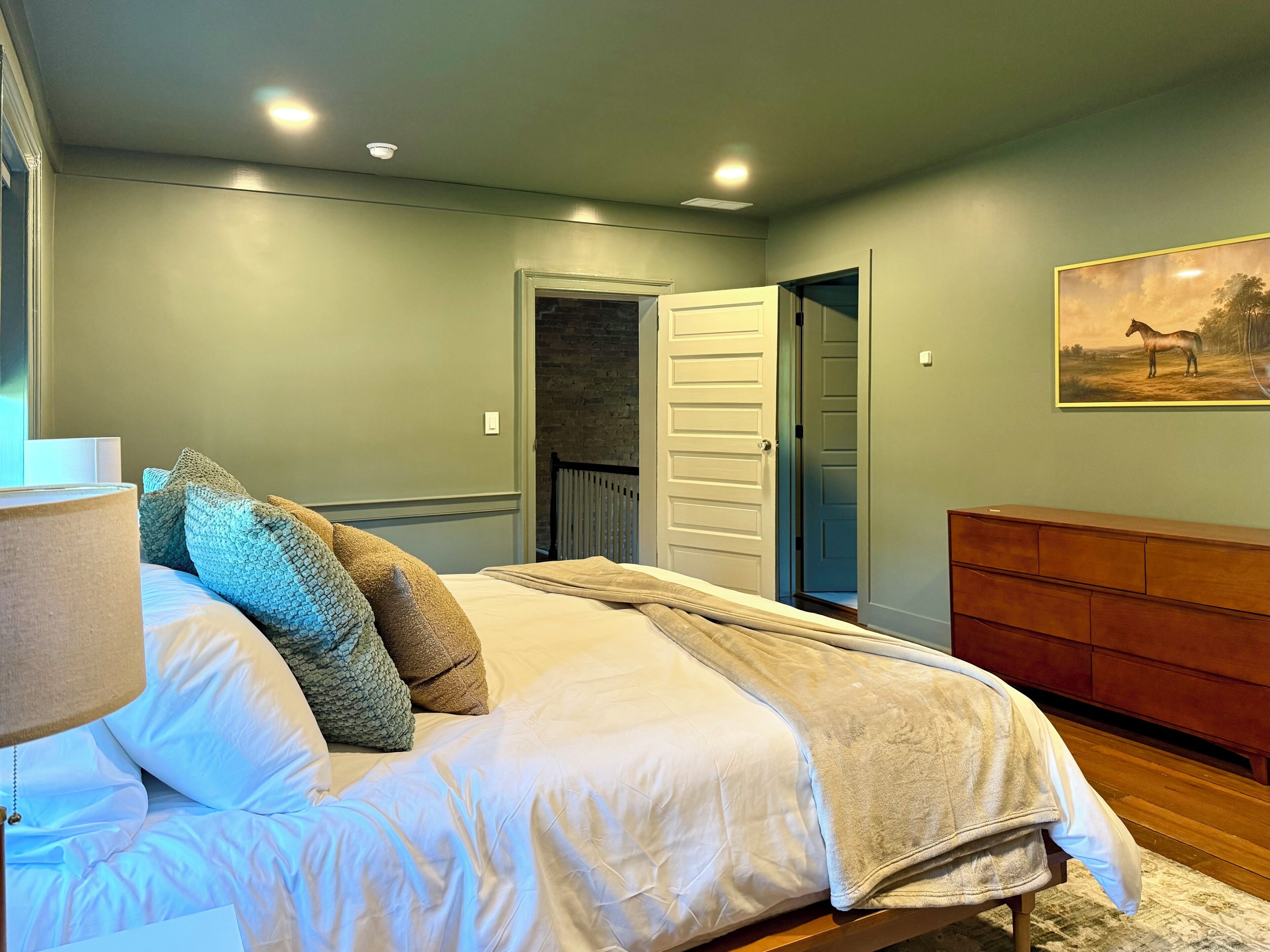A bedroom with a bed, a nightstand, a lamp, a door, and a painting on the wall.