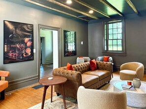 A modern living room with a brown leather sofa, a round wooden side table, and a beige armchair. There are two windows providing natural light and a large framed picture on the wall.