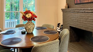 Uma sala de jantar com mesa redonda, cadeiras, um vaso de flores, lareira e janela com vista para árvores.