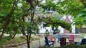 Um jardim com caminhos de pedra, mobiliário de exterior e um edifício branco com telhado vermelho ao fundo.