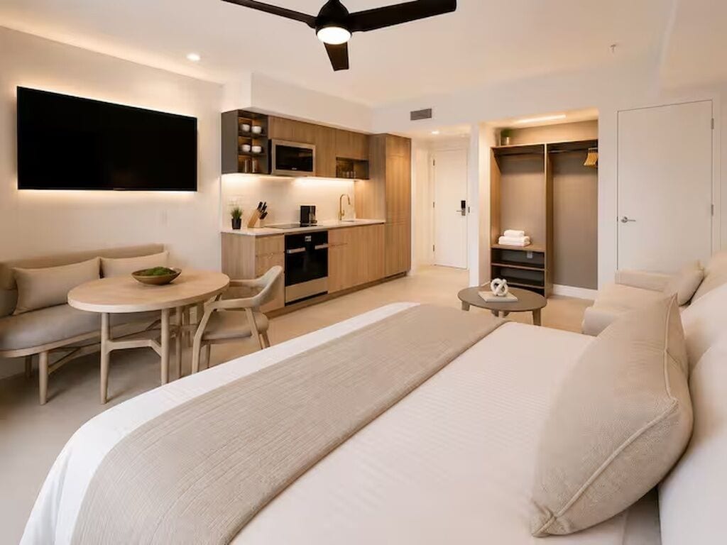 A modern hotel room with a large bed, a dining area, a kitchenette, and a walk-in closet.