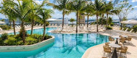 A swimming pool with lounge chairs and tables, surrounded by palm trees, with a view of a city skyline.
