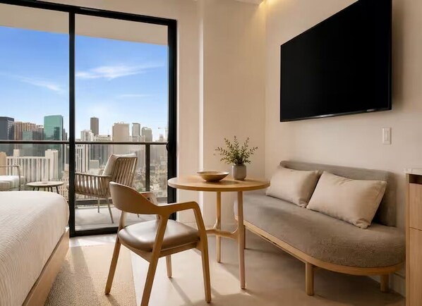 Habitación de hotel con cama, sofá, mesa de comedor con sillas, planta en maceta y televisor montado en la pared.