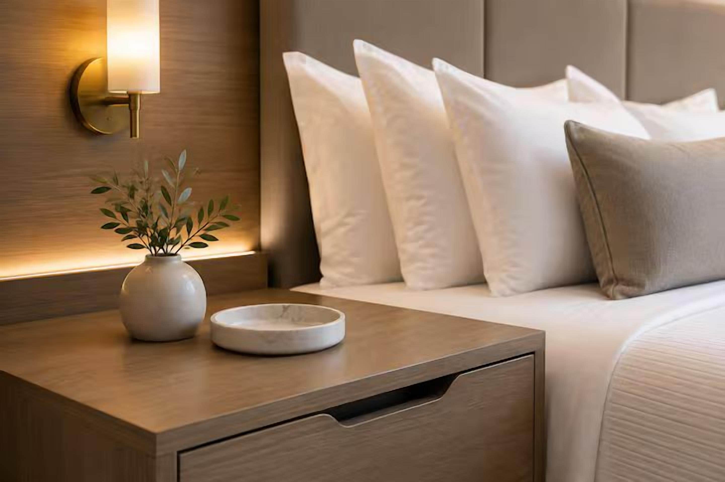 Un lit bien fait, avec une literie blanche, une table de chevet ornée d’un vase et d’un plateau, et un luminaire fixé au mur.