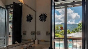 Ein Badezimmer mit einem großen Spiegel, einem Waschbecken und Blick auf ein Schwimmbad und Berge durch eine offene Tür.