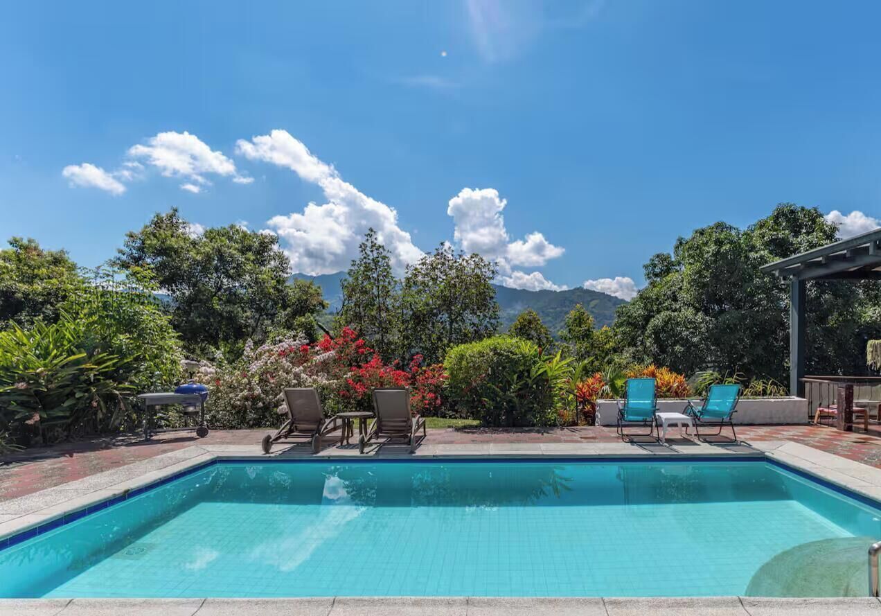 A rectangular swimming pool with clear blue water, surrounded by lounge chairs and potted plants, with a view of mountains in the background.