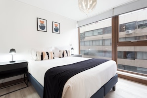 A modern bedroom with a large bed, bedside tables, a desk, and a view of the city through a large window. - Prime location in Las Condes | Just steps from the Manquehue Metro station (Las Condes)