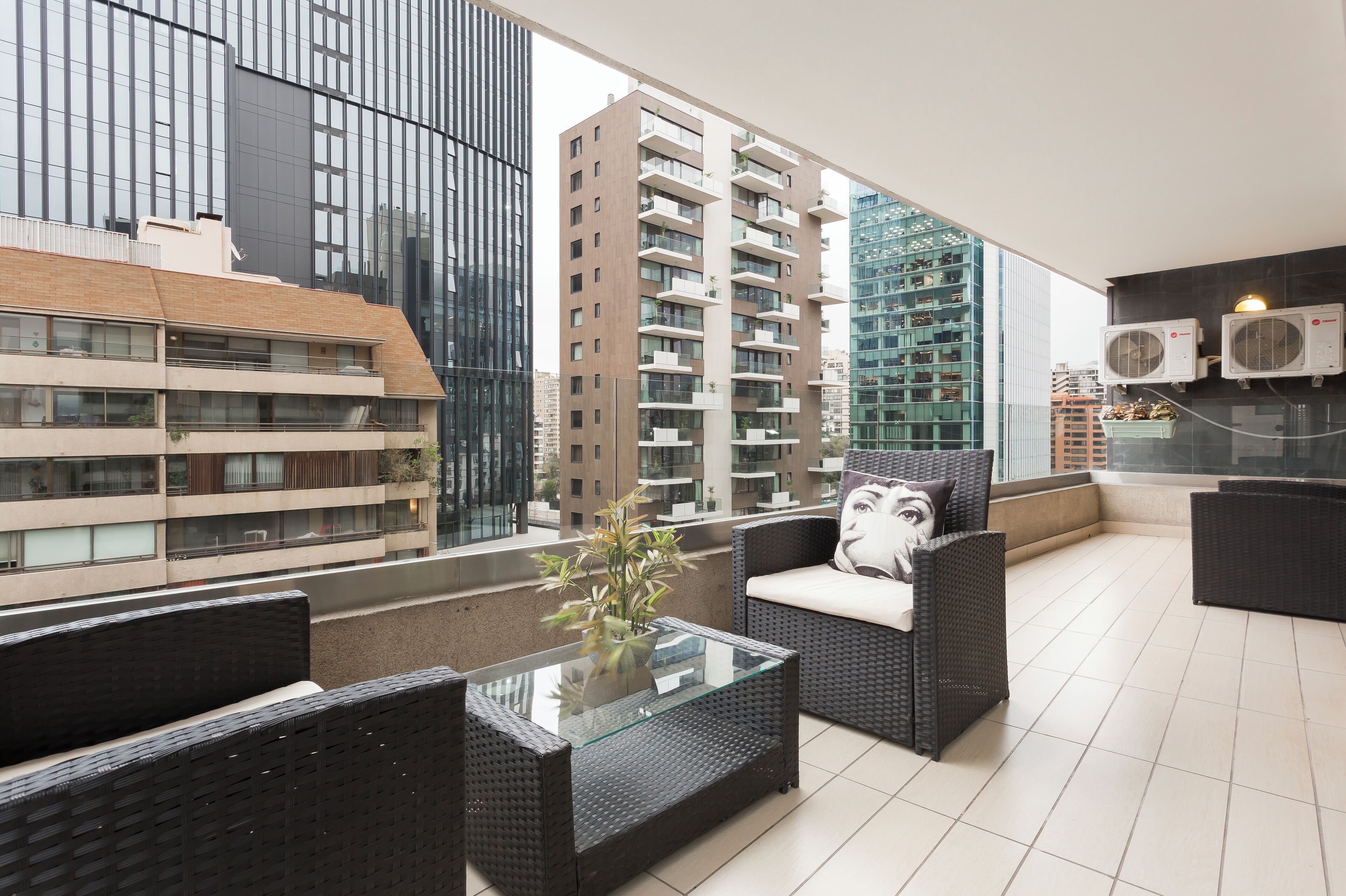 A modern balcony with wicker furniture, a glass table, and a view of surrounding buildings.