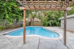 Ein Schwimmbecken mit einer hölzernen Pergola darüber, umgeben von einer Betonplattform und einem Holzzaun.
