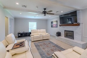 A modern living room with a fireplace, a ceiling fan, and a large window. - Energy Corridor 4BR | Near I-10 & Hwy 6 (Houston)