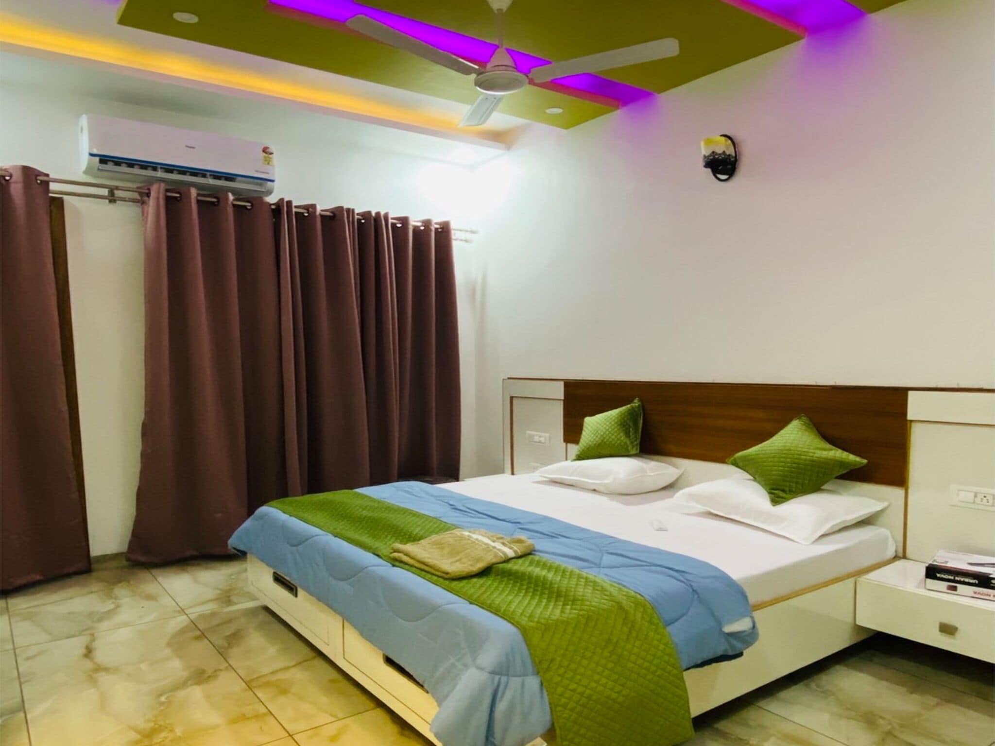 A hotel room with a bed, curtains, a ceiling fan, an air conditioning unit, and a bedside table with a book.