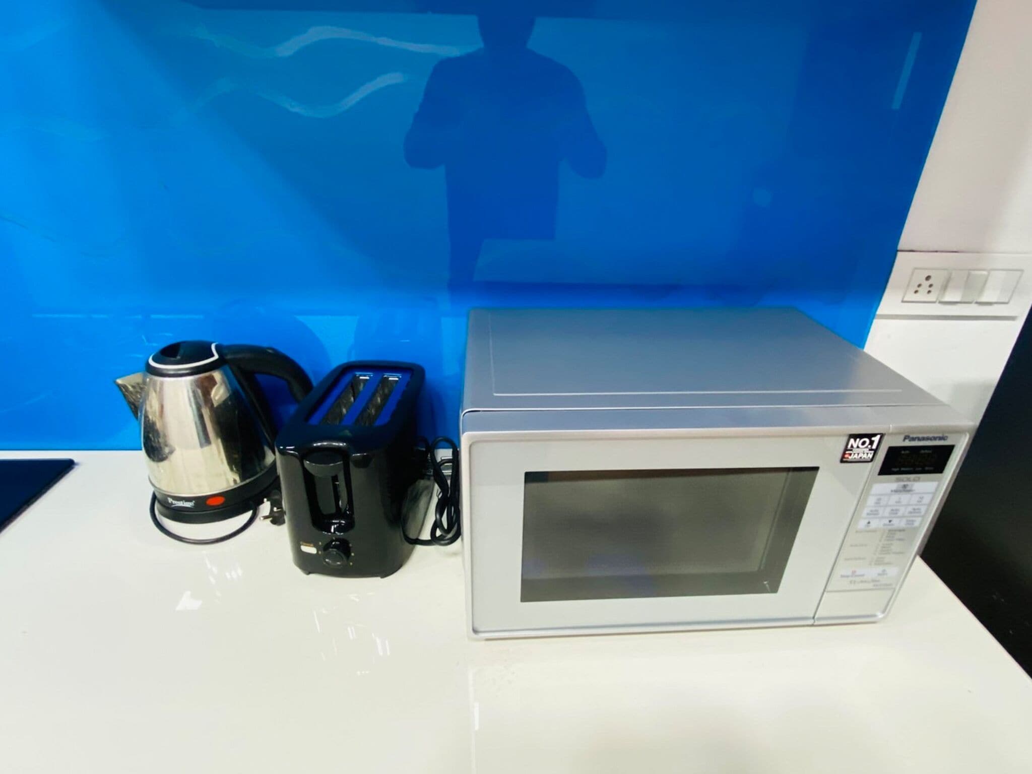 Un four à micro-ondes doté d’un écran numérique et d’un panneau de commande, une bouilloire électrique argentée et un grille-pain électrique noir, le tout posé sur un plan de travail blanc.