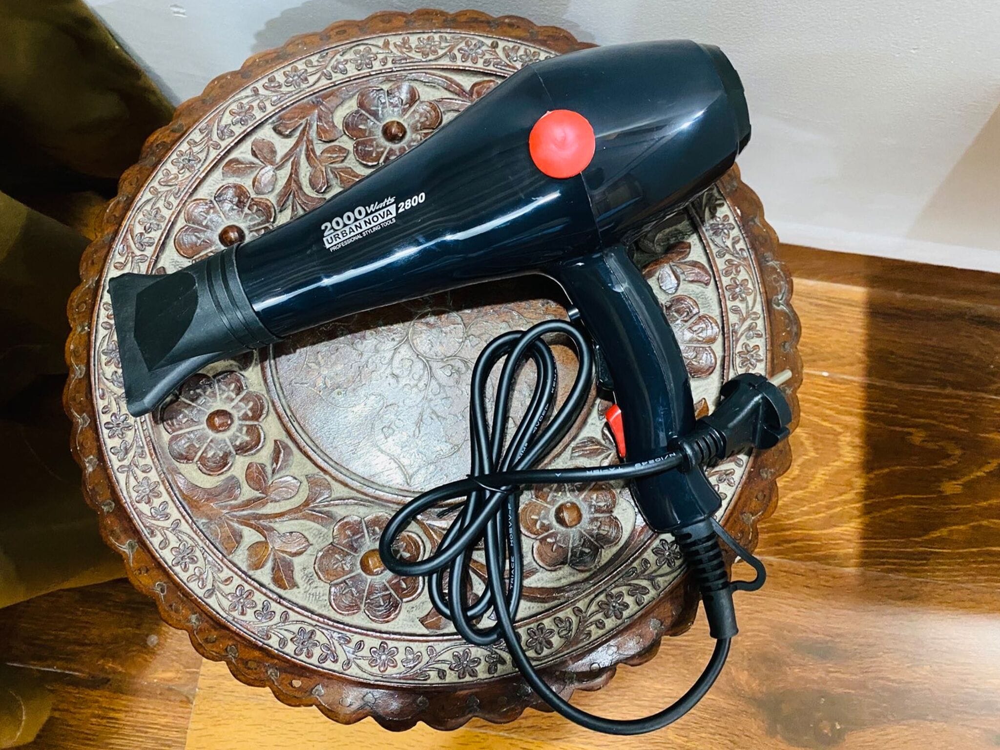 Un sèche-cheveux noir, doté d’un bouton rouge et d’un cordon, posé sur une assiette décorative.