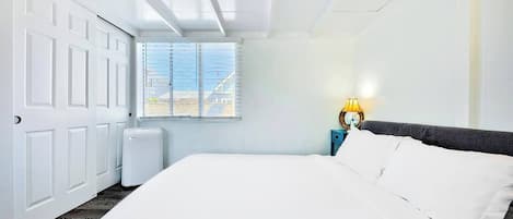 A hotel room with a large bed, a bedside table with a lamp, a window with blinds, and a white door.