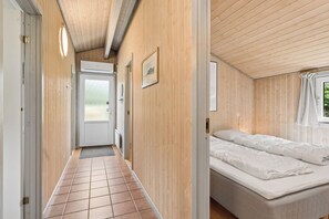 A narrow hallway with a door leading to a bedroom. The room has a bed with white bedding and a window with curtains. - Holiday Home in Jegum Ferieland (Oksbøl)