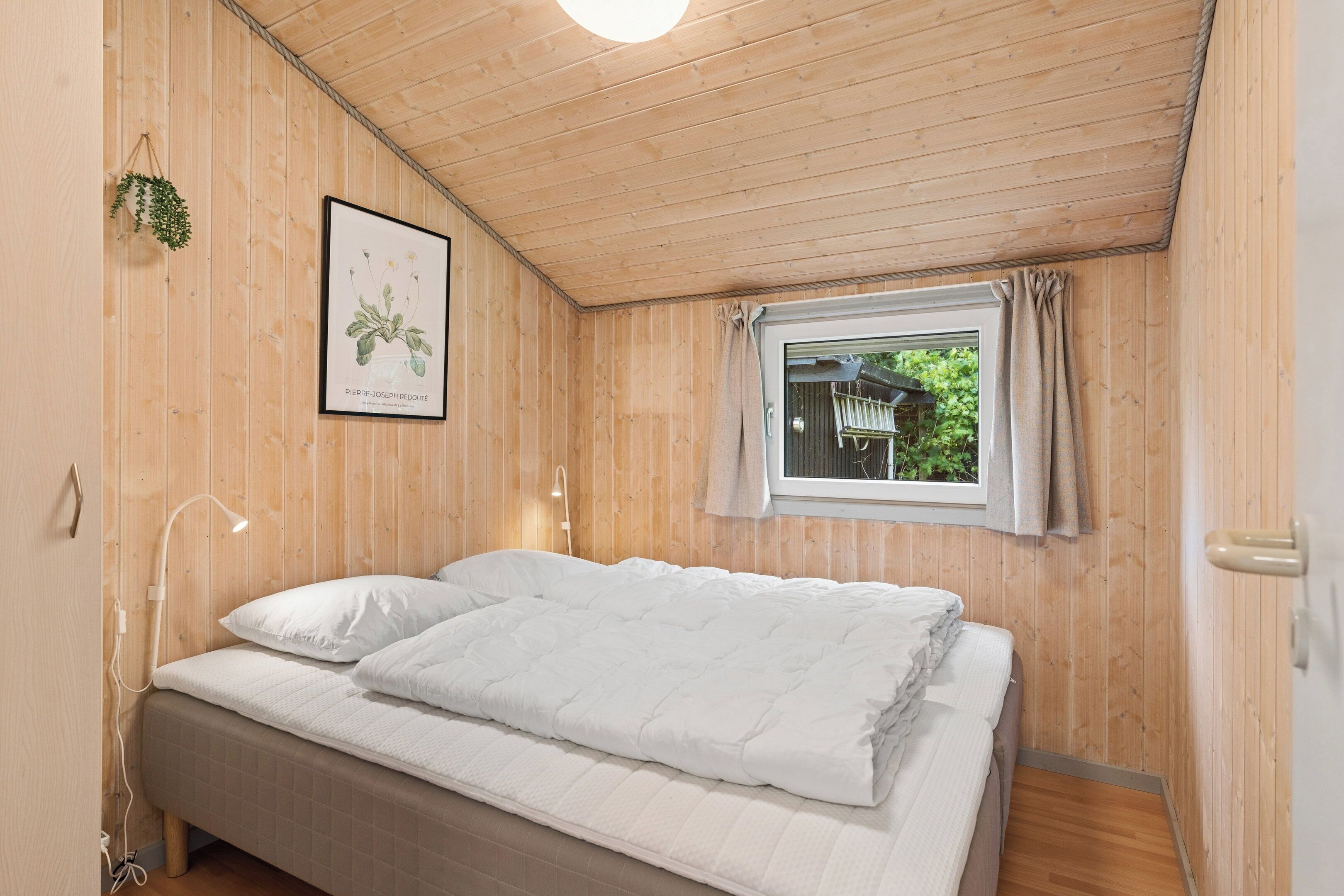Une chambre aux murs lambrissés, avec un lit, une illustration botanique encadrée et une fenêtre avec des rideaux.