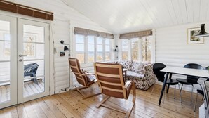 Een kamer met houten vloer, een bank, een eettafel en stoelen. Er zijn ramen met gordijnen, een deur en een lamp aan de muur.