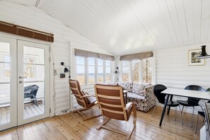 A room with a wooden floor, a sofa, a dining table, and chairs. There are windows with curtains, a door, and a lamp on the wall. - Authentic holiday home with Baltic Sea view (Allinge-Sandvig)