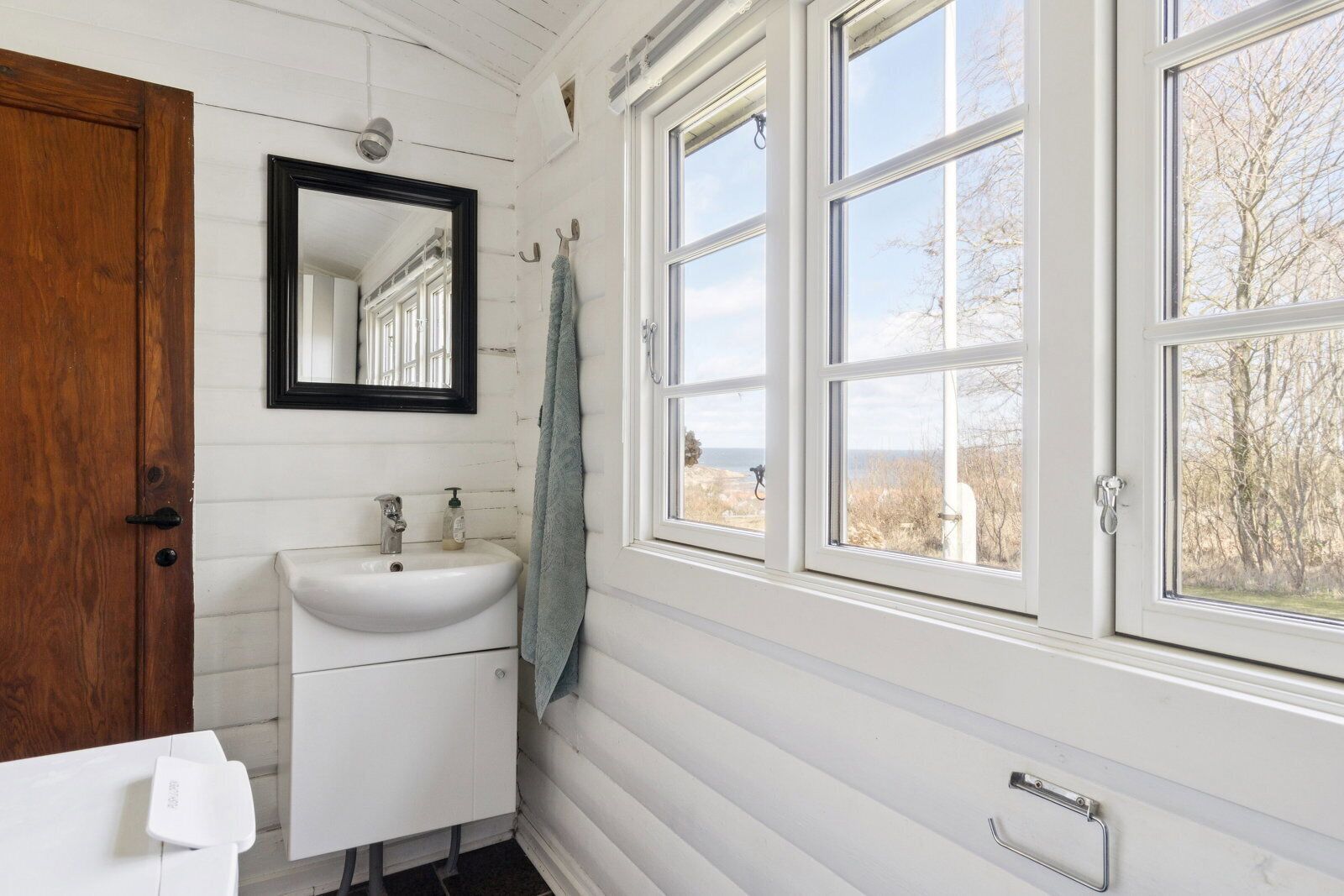 Ein Badezimmer mit weißem Waschbecken, einem Spiegel und einem Fenster mit Blick ins Freie.