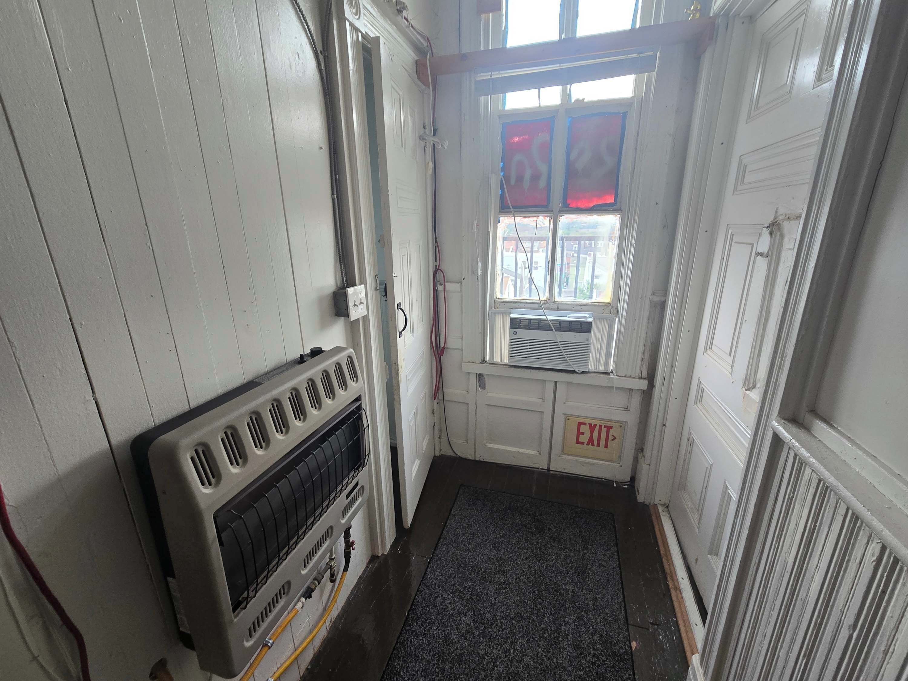 Un couloir étroit avec un radiateur, un panneau indiquant la sortie et une fenêtre dotée de vitres teintées en rouge.