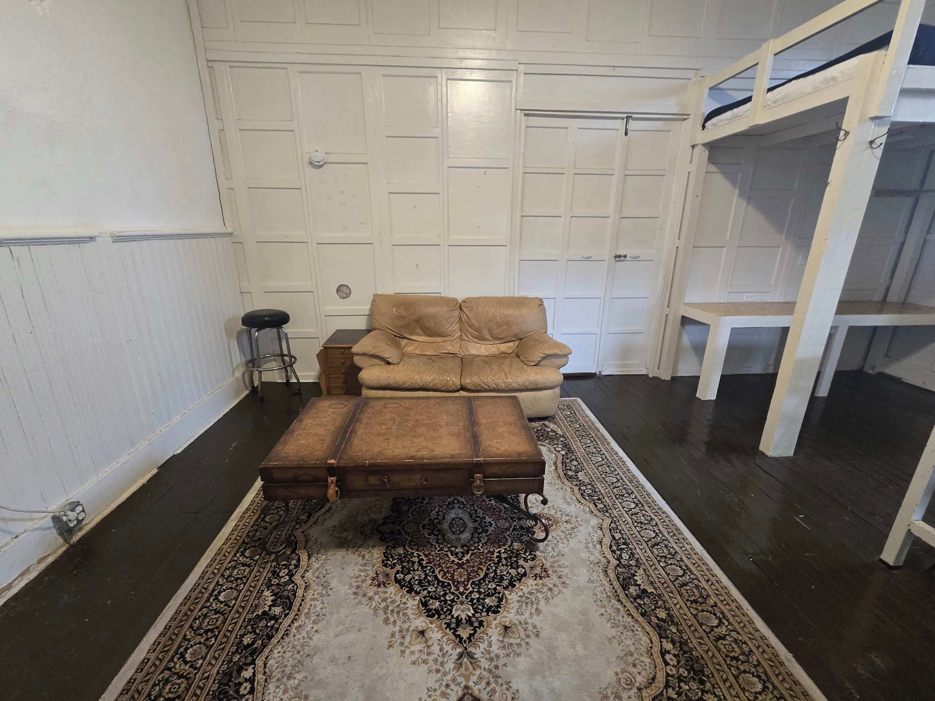 Une pièce comprenant un fauteuil beige, un pouf en bois, un tapis à motifs, un petit tabouret et une étagère blanche adossée à un mur blanc à panneaux.