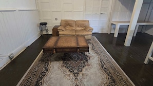 Ett rum med en beige fåtölj, en träpall, en mönstrad matta, en liten pall och en vit hylla som står mot en vit panelvägg.