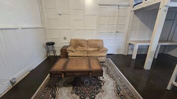 Une pièce comprenant un fauteuil beige, un pouf en bois, un tapis à motifs, un petit tabouret et une étagère blanche adossée à un mur blanc à panneaux.