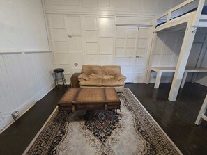 Ein Zimmer mit einem beige-farbenen Sessel, einem hölzernen Hocker, einem gemusterten Teppich, einem kleinen Stuhl und einer weißen Regaleinheit an einer weiß gefliesten Wand.