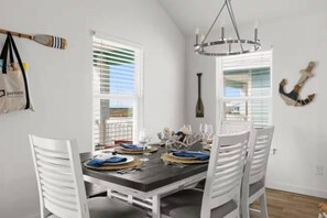 A dining area with a dark wooden table, white chairs, and a chandelier. - Heated Pool Open All Year | 4BR Sleeps 10 | Firepit, Games & Fenced Yard (Port Aransas)