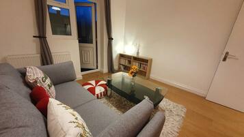 A living room with a grey sofa, a glass coffee table, a bookshelf, and a door.