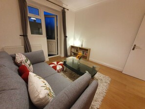 A living room with a grey sofa, a glass coffee table, a bookshelf, and a door. - Cosy and very Family-sized with Game and TV rooms and relaxing  & Friendly (London)