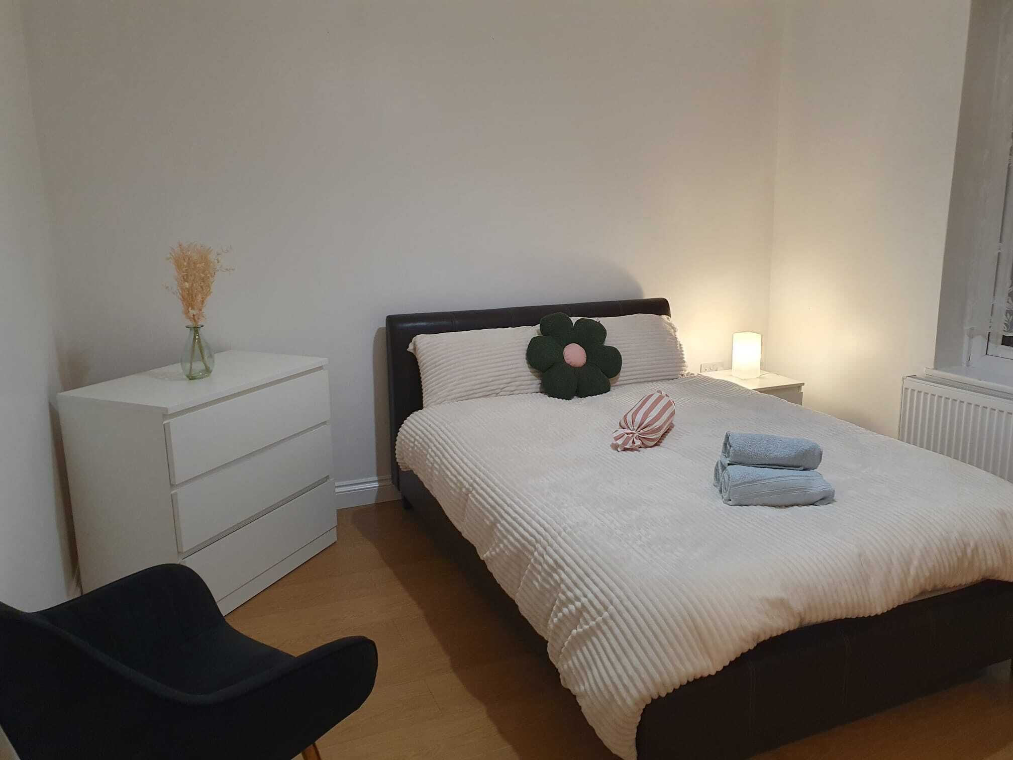 Een slaapkamer met een bed, nachtkastje, lamp, stoel en kledingkast.
