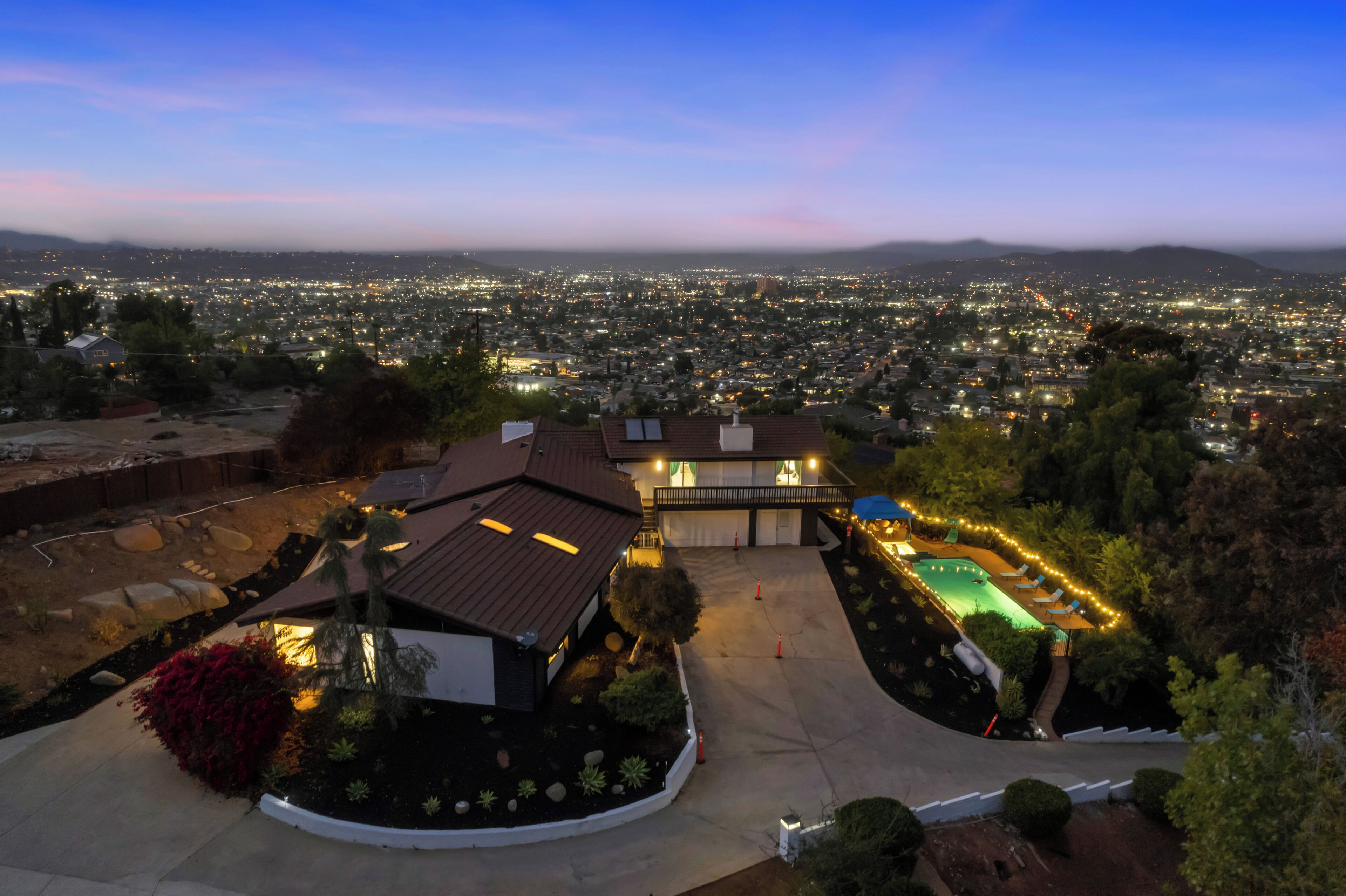 A residential property with a swimming pool, a covered patio, and a view of a cityscape at dusk.