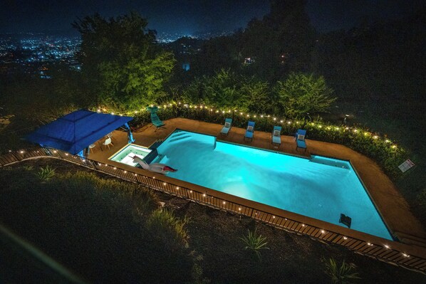 A rectangular swimming pool with a blue umbrella and lounge chairs around it, surrounded by trees and string lights.