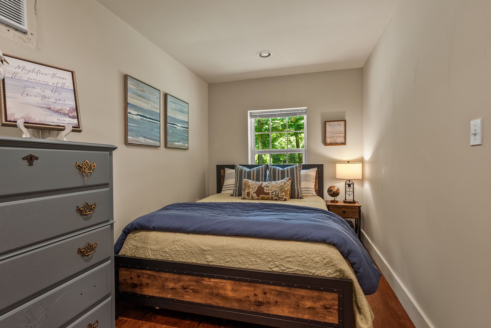 A bedroom with a bed, dresser, and a window with a view of greenery.