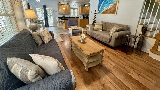 A spacious living room with a dark-colored sofa, a wooden coffee table, and a beige sofa. A kitchen is visible in the background with wooden cabinets and a dining area. - Ocean Lakes Beach House-Golf Cart Included-Fire Pit-King Suite-Linens (Myrtle Beach)