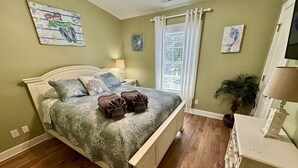 A bedroom with a bed, bedside tables, a lamp, a potted plant, and artwork on the walls. - Ocean Lakes Beach House-Golf Cart Included-Fire Pit-King Suite-Linens (Myrtle Beach)