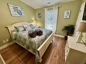 A bedroom with a bed, bedside tables, a lamp, a potted plant, and artwork on the walls. - Ocean Lakes Beach House-Golf Cart Included-Fire Pit-King Suite-Linens (Myrtle Beach)