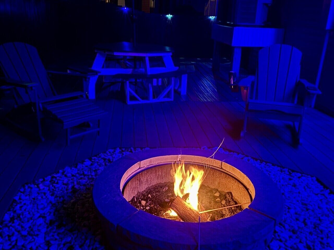 Ein runder Feuerkorb mit Flammen, umgeben von einer Holzterrasse mit Adirondack-Stühlen und einem Tisch.