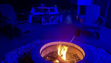 A circular fire pit with flames, surrounded by a wooden deck with Adirondack chairs and a table.
